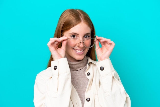 Jeune femme rousse isolée sur fond bleu avec des lunettes avec une expression heureuse