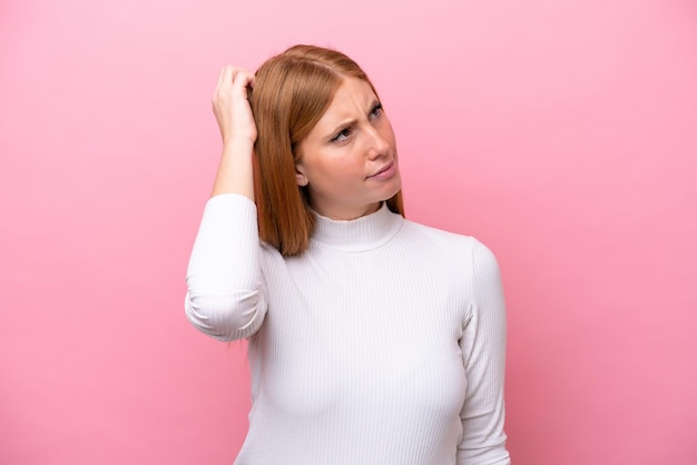 Jeune femme rousse isolée sur fond rose ayant des doutes en se grattant la tête