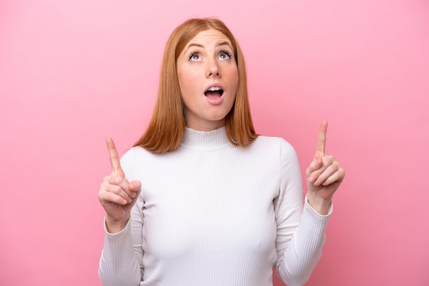 Jeune femme rousse isolée sur fond rose surprise et pointant vers le haut