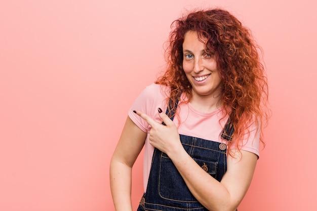 Jeune femme rousse jolie gingembre portant une salopette jeans souriant et pointant de côté, montrant quelque chose à l'espace vide.