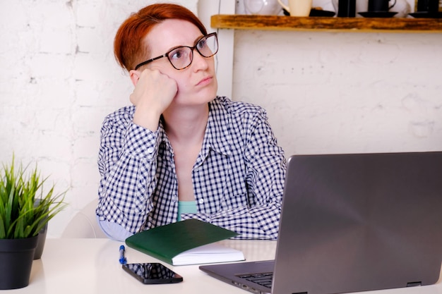 Une jeune femme rousse à lunettes est assise devant un écran d'ordinateur portable, appuyant sa tête sur sa main et ayant l'air ennuyée sur le côté. Le concept d'utilisation improductive du temps de travail lors des réunions en ligne.