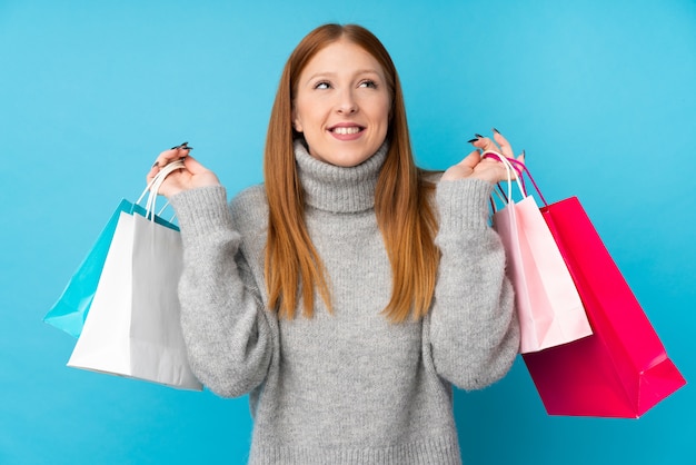 Jeune femme rousse sur mur bleu isolé tenant des sacs à provisions et souriant