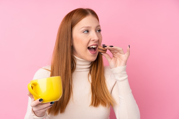 Jeune Femme Rousse Sur Un Mur Rose Isolé Tenant Des Macarons Français Colorés Et Une Tasse De Lait