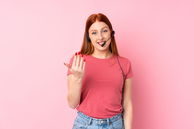 Jeune femme rousse sur mur rose isolé travaillant avec casque faisant geste à venir