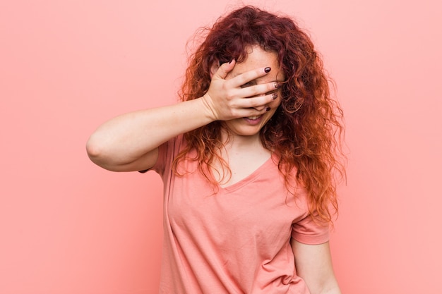 Jeune femme rousse naturelle et authentique clignote des yeux à la caméra à travers les doigts, embarrassée couvrant le visage.