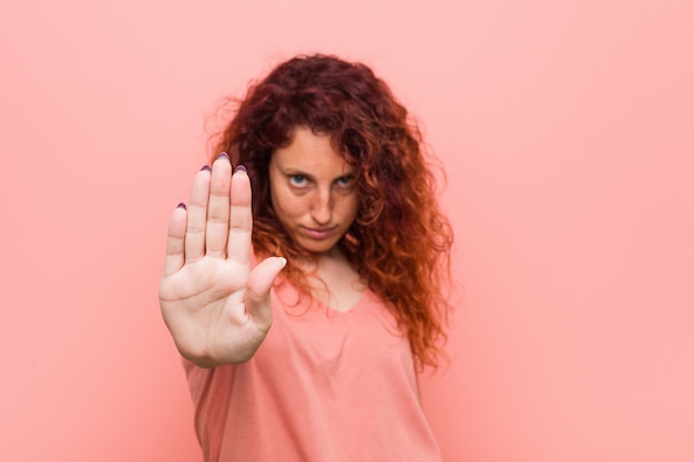 Jeune femme rousse naturelle et authentique debout avec la main tendue montrant le panneau d'arrêt