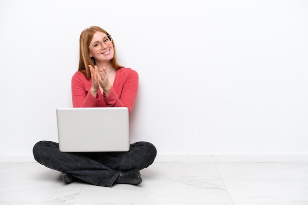 Jeune femme rousse avec un ordinateur portable assis sur le sol isolé sur fond blanc applaudissant après présentation lors d'une conférence