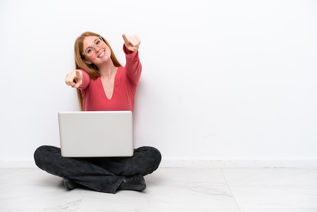 Jeune femme rousse avec un ordinateur portable assis sur le sol isolé sur fond blanc pointe le doigt vers vous en souriant