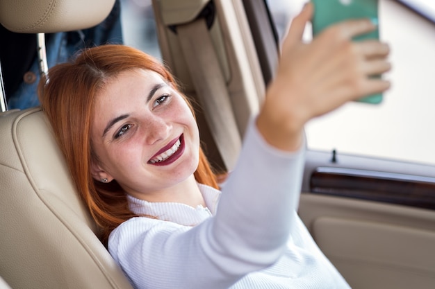 Jeune femme rousse pilote prenant selfies avec son téléphone portable assis derrière le volant de la voiture dans les embouteillages aux heures de pointe.