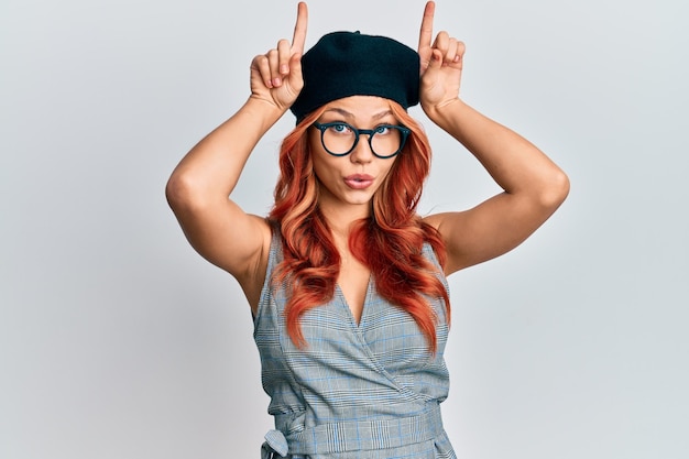 Une jeune femme rousse portant un look français à la mode avec un béret faisant un geste drôle avec le doigt sur la tête comme des cornes de taureau