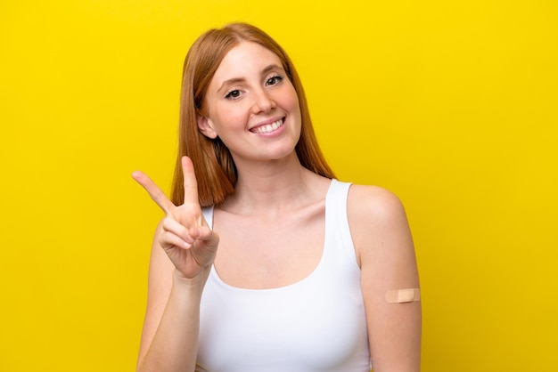 Jeune femme rousse portant un pansement isolé sur fond jaune souriant et montrant le signe de la victoire