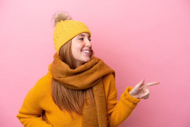 Jeune femme rousse portant une veste d'hiver isolée sur fond rose pointant le doigt sur le côté et présentant un produit