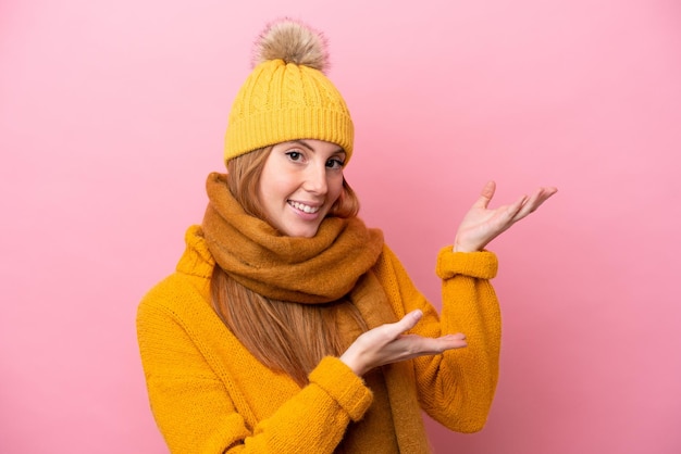 Jeune femme rousse portant une veste d'hiver isolée sur fond rose tendant les mains sur le côté pour inviter à venir