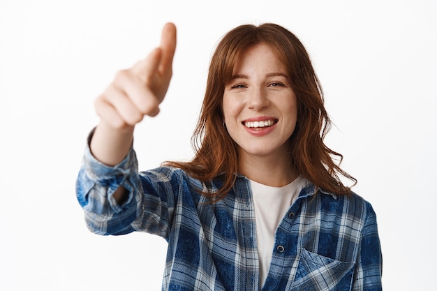 Jeune femme rousse riant, pointant du doigt l'écran de télévision et souriant heureuse sur blanc