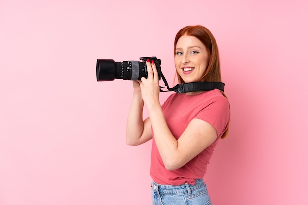 Jeune femme rousse sur rose isolé avec un appareil photo professionnel