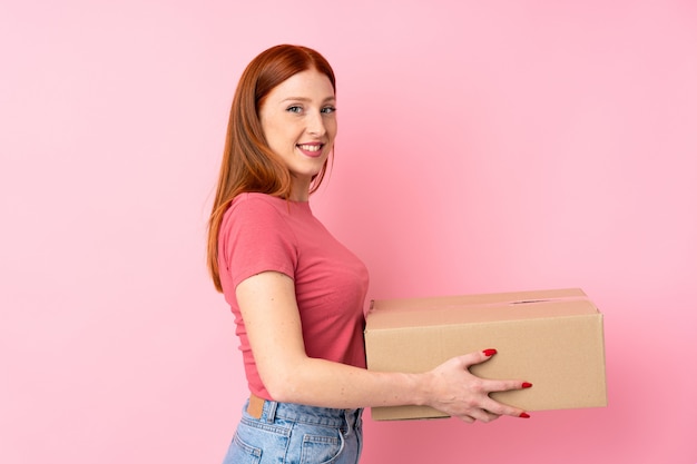 Jeune femme rousse sur rose isolé tenant une boîte pour la déplacer vers un autre site