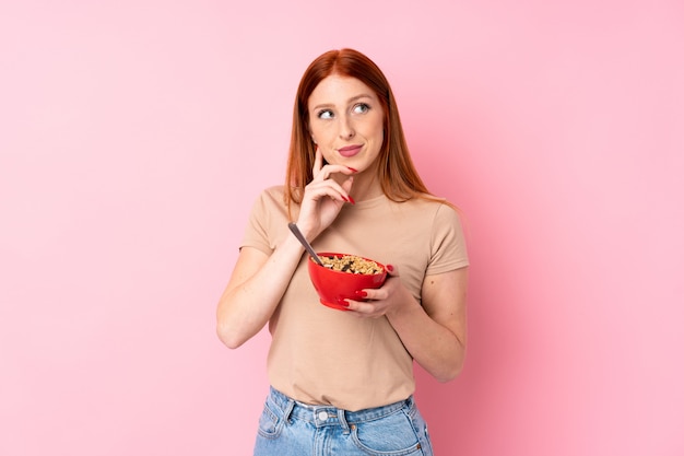 Jeune femme rousse sur rose isolé tenant un bol de céréales et de penser