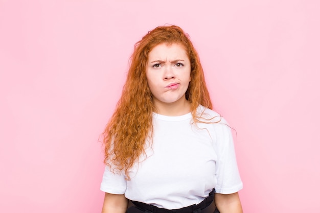 Jeune femme rousse se sentant confuse et douteuse, se demandant ou essayant de choisir ou de prendre une décision sur le mur rose