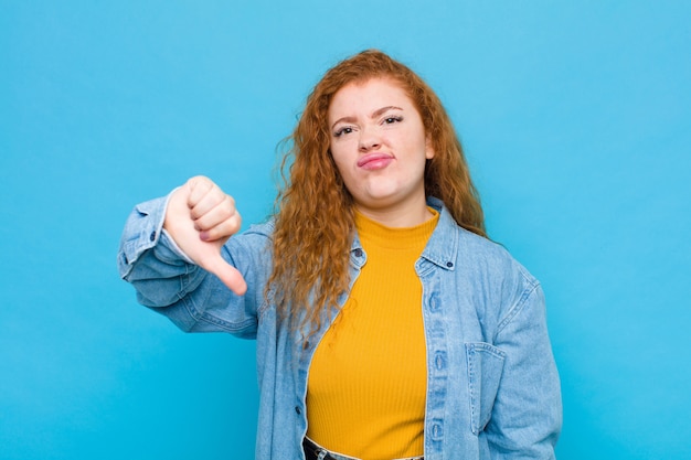 Jeune femme rousse se sentant en croix, en colère, ennuyée, déçue ou mécontente, montrant les pouces vers le bas avec un regard sérieux sur le mur bleu