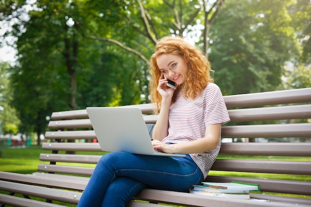 Jeune femme rousse souriante parlant sur smartphone tenant un ordinateur portable sur le genou assis à l'extérieur sur le banc dans le parc