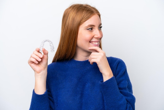 Jeune femme rousse tenant des accolades invisibles isolées sur fond blanc en pensant à une idée et en regardant de côté