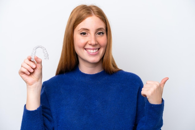 Jeune femme rousse tenant des accolades invisibles isolées sur fond blanc pointant vers le côté pour présenter un produit