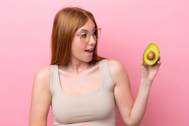 Jeune femme rousse tenant un avocat isolé sur fond rose avec une expression surprise tout en regardant de côté