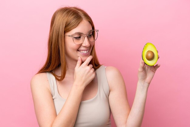 Jeune femme rousse tenant un avocat isolé sur fond rose regardant sur le côté et souriant