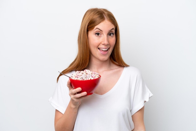 Jeune femme rousse tenant un bol de céréales isolé sur fond blanc avec une expression faciale surprise et choquée