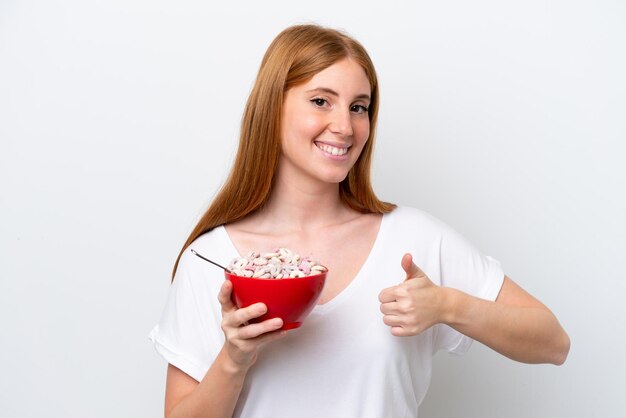 Jeune femme rousse tenant un bol de céréales isolé sur fond blanc avec les pouces vers le haut parce que quelque chose de bien s'est produit