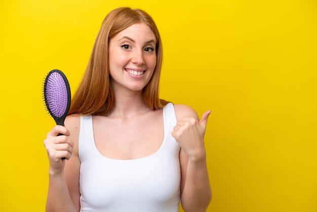 Jeune femme rousse tenant une brosse à cheveux isolée sur fond blanc pointant vers le côté pour présenter un produit