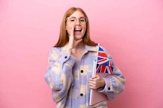 Jeune femme rousse tenant un drapeau du Royaume-Uni isolé sur fond rose criant avec la bouche grande ouverte