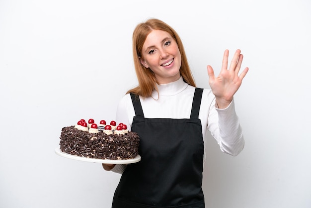 Jeune femme rousse tenant un gâteau d'anniversaire isolé sur fond blanc en comptant cinq avec les doigts