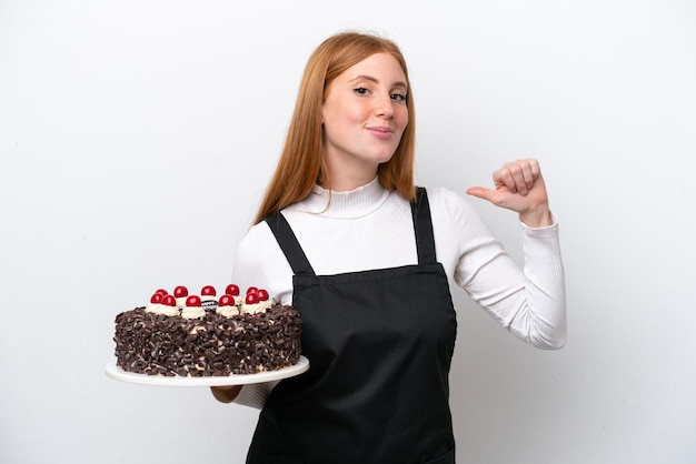 Jeune femme rousse tenant un gâteau d'anniversaire isolé sur fond blanc fier et satisfait