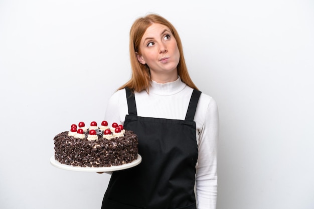 Jeune femme rousse tenant un gâteau d'anniversaire isolé sur fond blanc et levant les yeux