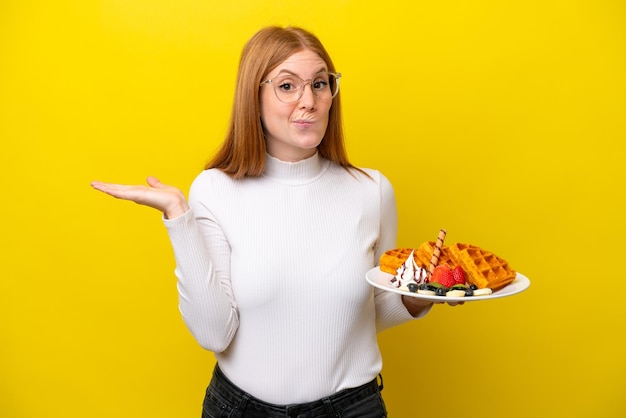 Jeune femme rousse tenant des gaufres isolées sur fond jaune ayant des doutes tout en levant les mains