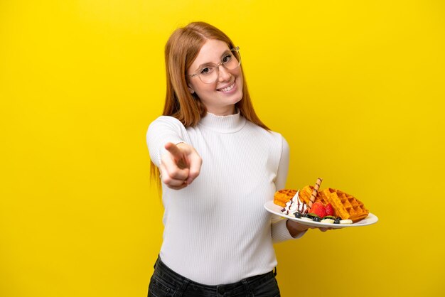 Jeune femme rousse tenant des gaufres isolées sur fond jaune pointant vers l'avant avec une expression heureuse