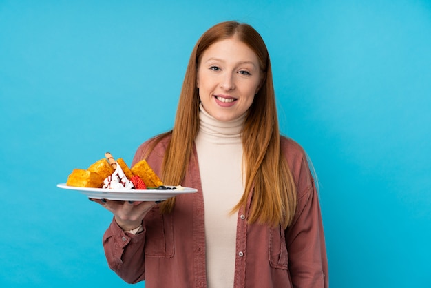 Jeune femme rousse tenant des gaufres sur un mur isolé, souriant beaucoup