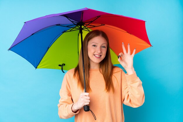 Jeune femme rousse tenant un parapluie sur un mur isolé montrant un signe ok avec les doigts