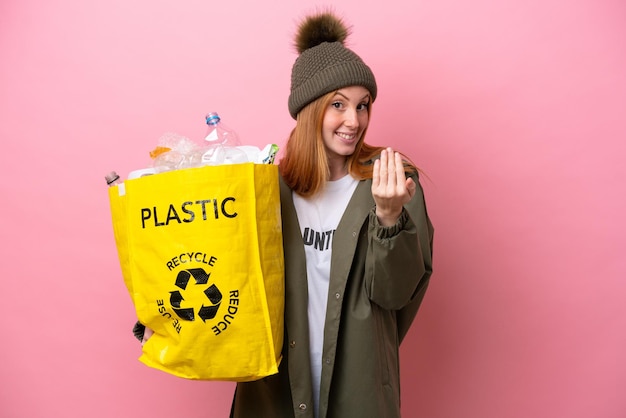 Jeune femme rousse tenant un sac plein de bouteilles en plastique à recycler isolé sur fond rose invitant à venir avec la main Heureux que vous soyez venu