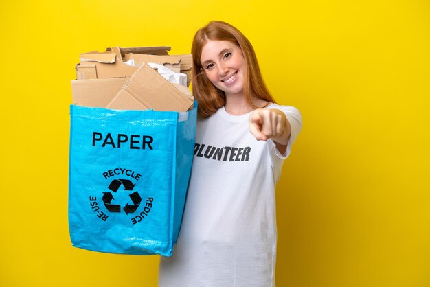 Jeune femme rousse tenant un sac de recyclage plein de papier à recycler isolé sur fond jaune pointant vers l'avant avec une expression heureuse