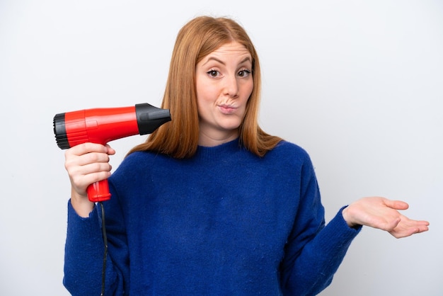 Photo jeune femme rousse tenant un sèche-cheveux isolé sur fond blanc faisant un geste de doute tout en soulevant les épaules