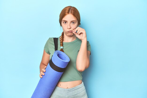 Jeune femme rousse tenant un tapis de yoga en studio avec les doigts sur les lèvres gardant un secret