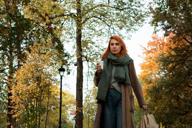 Jeune femme rousse en tenue d'automne élégante avec sac à provisions écologique tasse de café marchant dans le parc