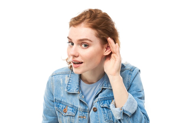 Jeune femme rousse en tenue décontractée souriante et tenant la main près de l'oreille en écoutant attentivement en entendant attentivement isolé sur fond blanc