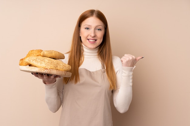 Jeune femme rousse en uniforme de chef. Boulanger tenant une table avec plusieurs pains pointant vers le côté pour présenter un produit