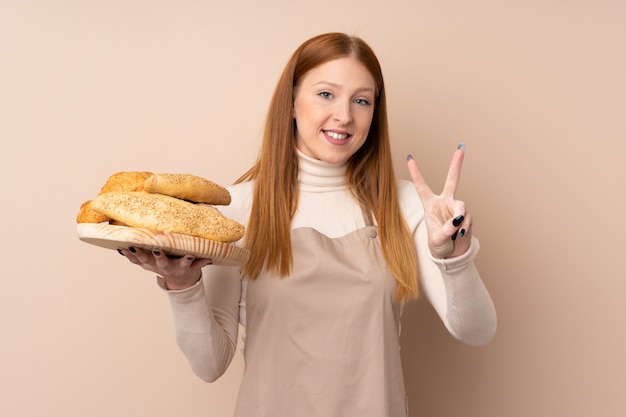 Jeune femme rousse en uniforme de chef. Femme boulanger tenant une table avec plusieurs pains souriant et montrant le signe de la victoire