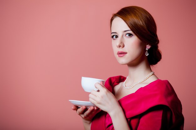 Jeune femme rousse en vêtements d'époque victorienne rouge avec tasse de thé sur rose