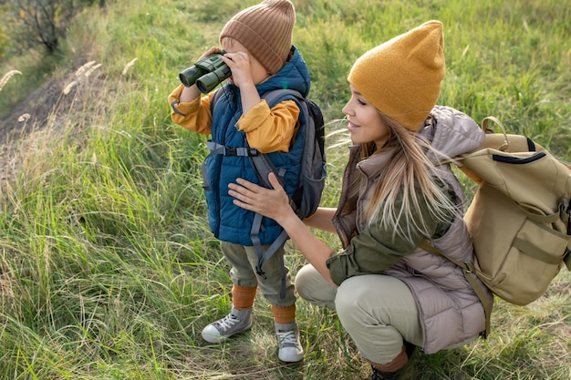 Jeune femme routarde en tenue décontractée chaude accroupie près de son adorable petit fils avec des jumelles lors d'un week-end dans un environnement naturel