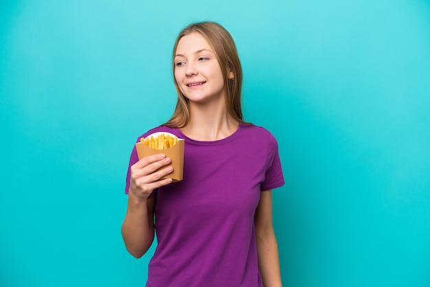 Jeune femme russe attrapant des frites isolées sur fond bleu regardant sur le côté et souriant